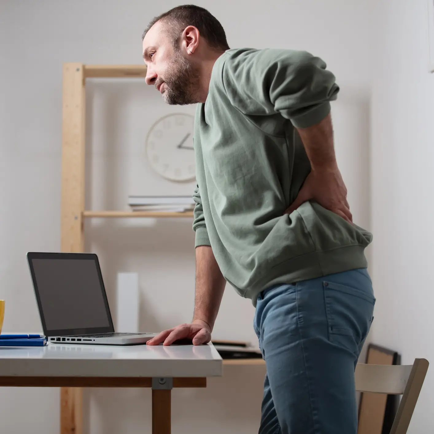 medelåldersman i grön sweatshirt och blå jeans står vid skrivbord och har ont i ryggen